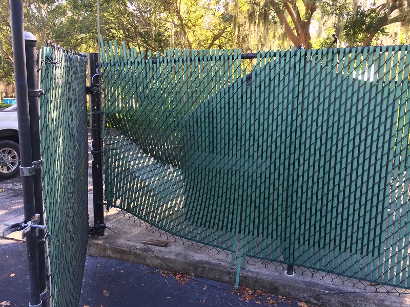 Damaged commercial dumpster enclosure chain link fence with green privacy slats requiring repair in Orlando, FL.